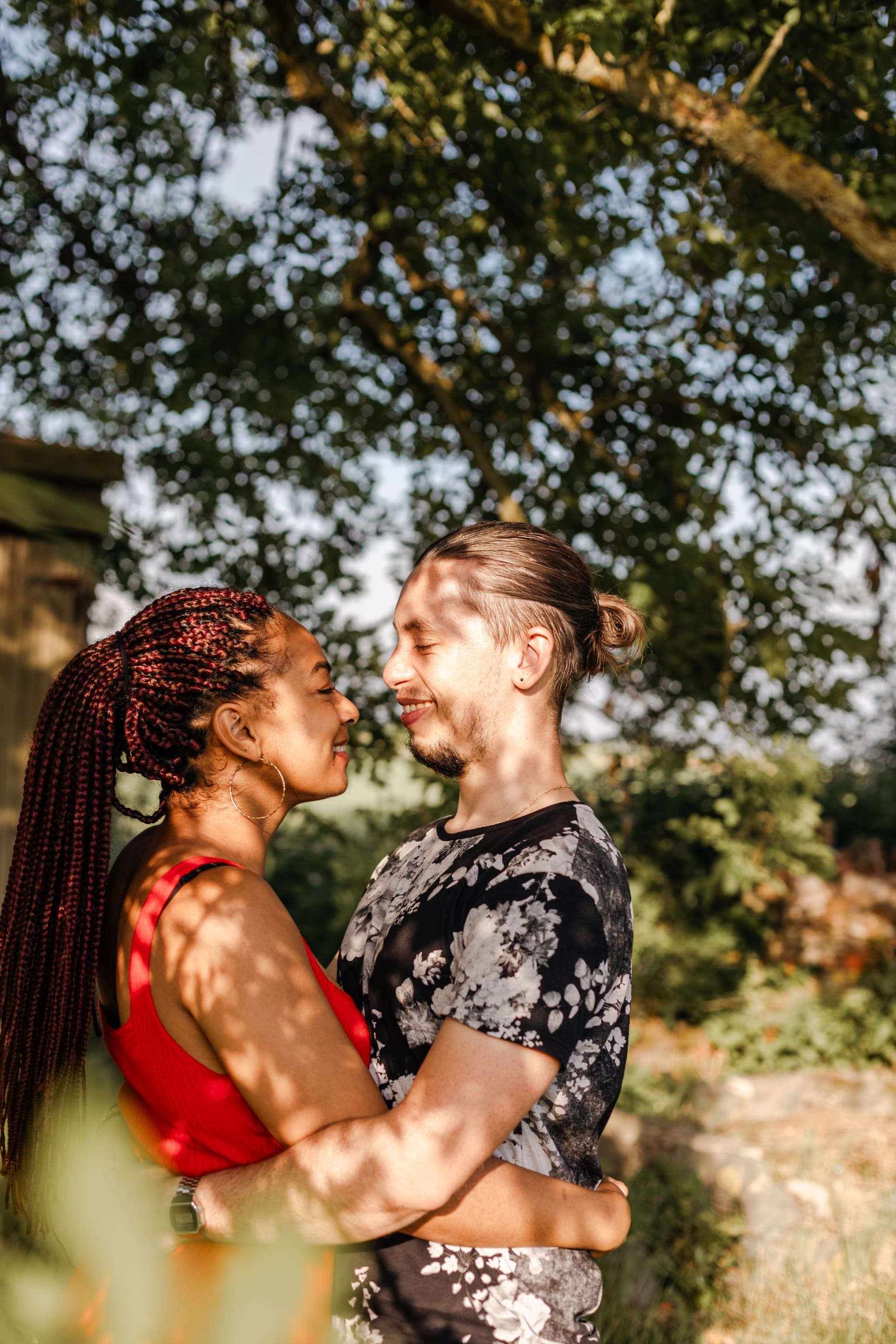 Couple enlacés nez à nez sous un arbre lumière filtrée femme aux tresses rouges — séance couple intime été
