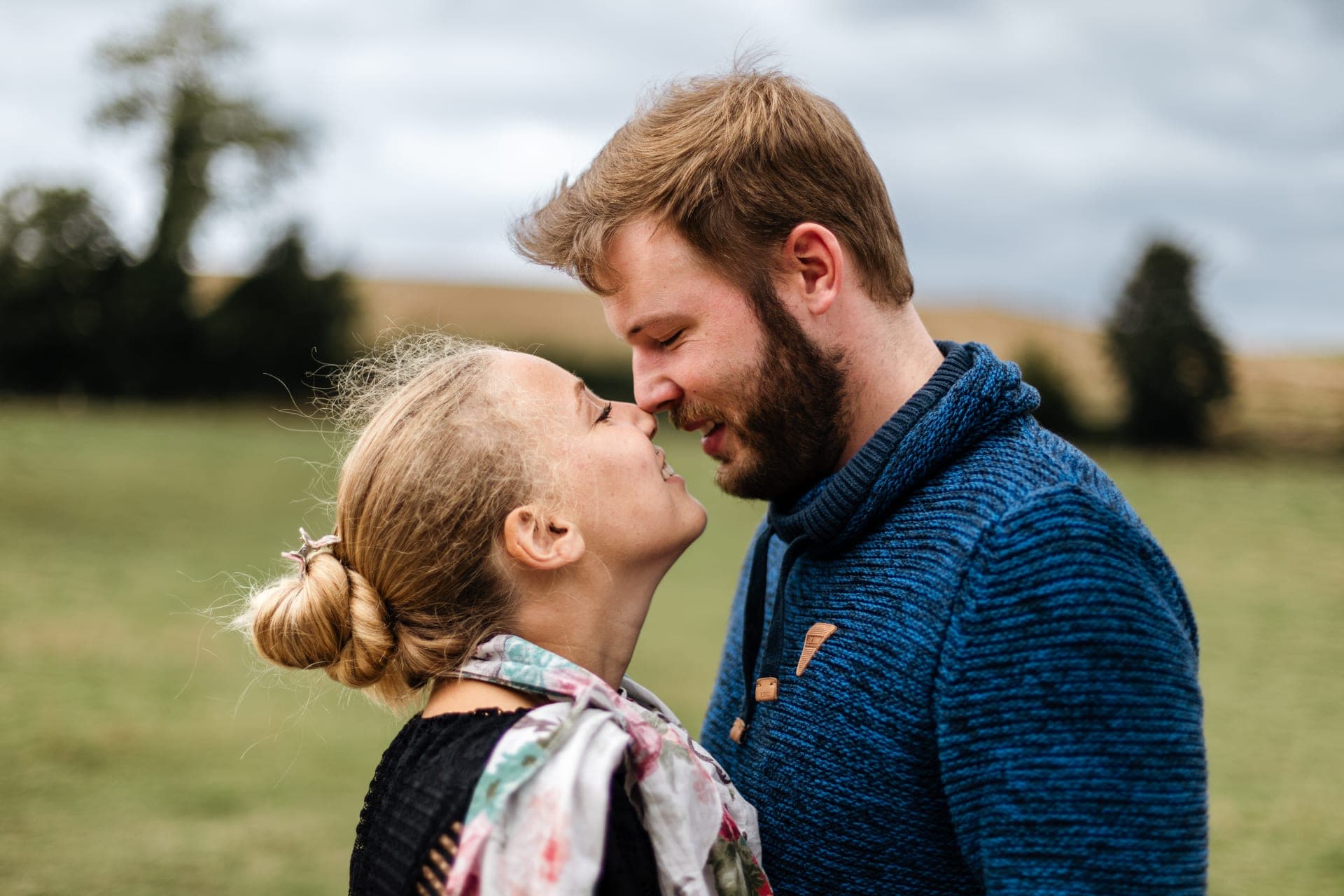 Couple nez à nez sourire complice dans un champ pull bleu marine ciel nuageux — séance couple campagne Aurore Clercq Photographie Brabant Wallon