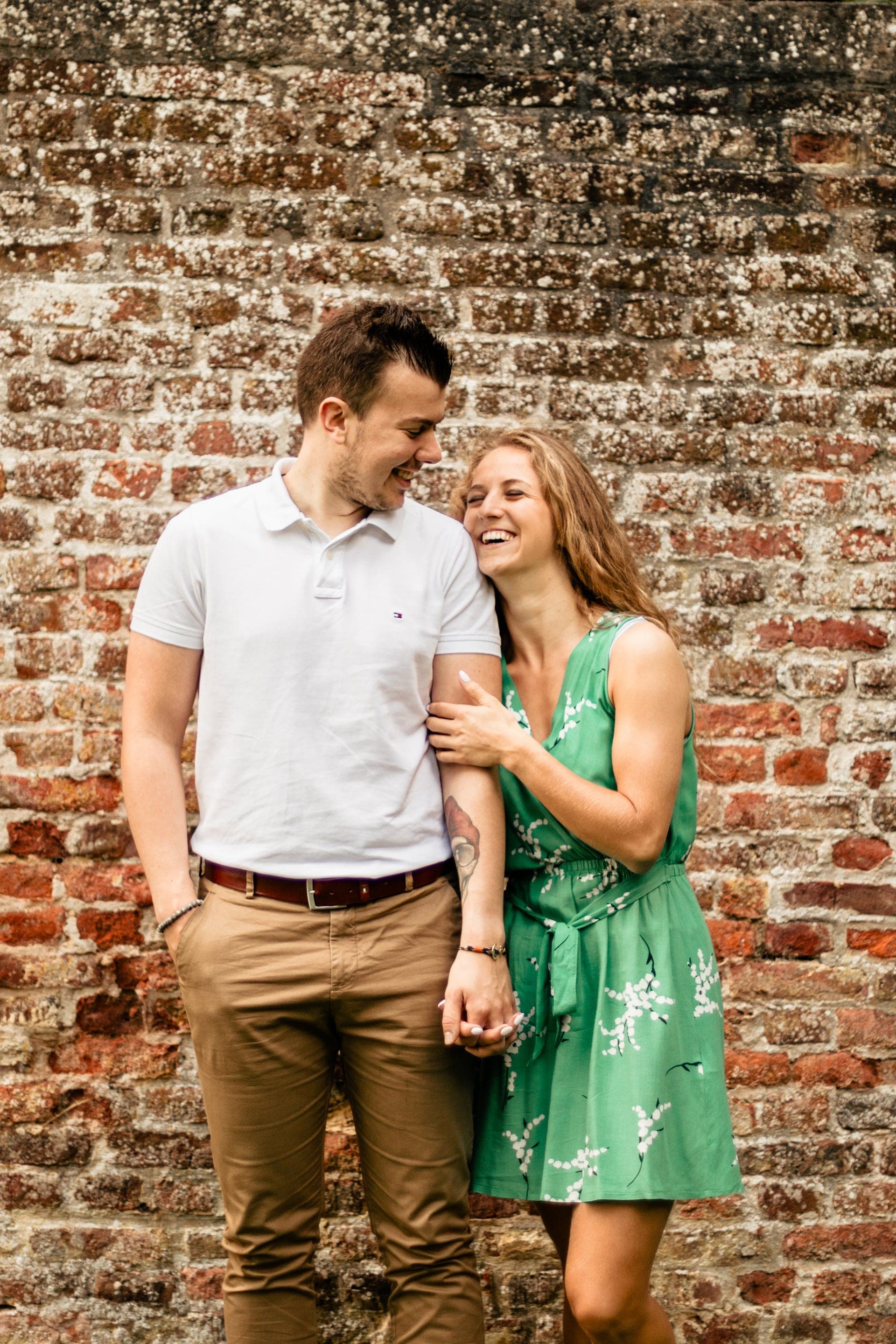 Couple en tenues harmonisées robe verte fleurie et polo blanc fou rire complice devant un mur de briques — exemple de tenues pour séance photo en Brabant Wallon