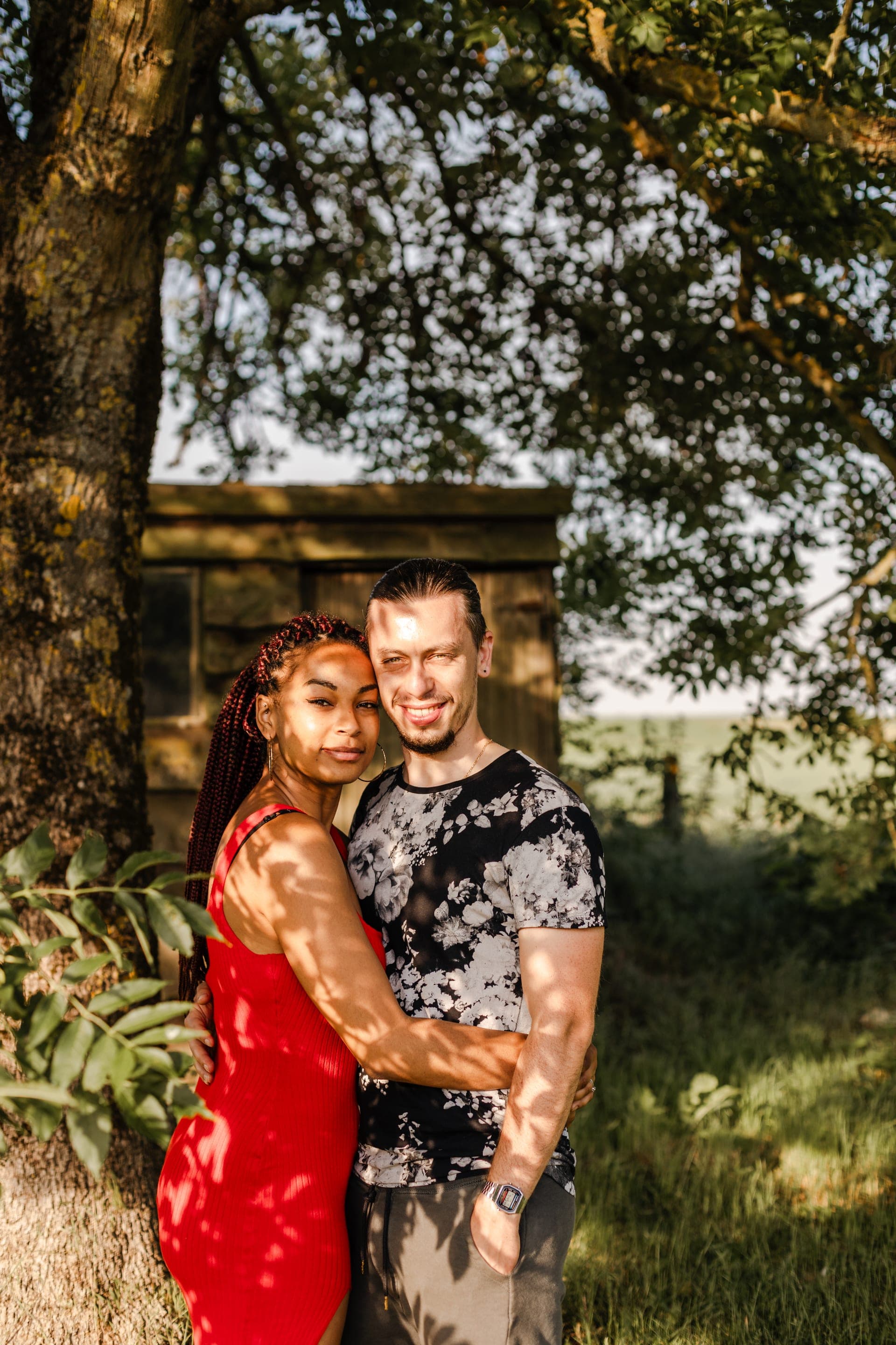 Couple enlacés sous un arbre devant une cabane en bois robe rouge tresses lumière dorée — séance couple été golden hour Aurore Clercq Photographie Brabant Wallon