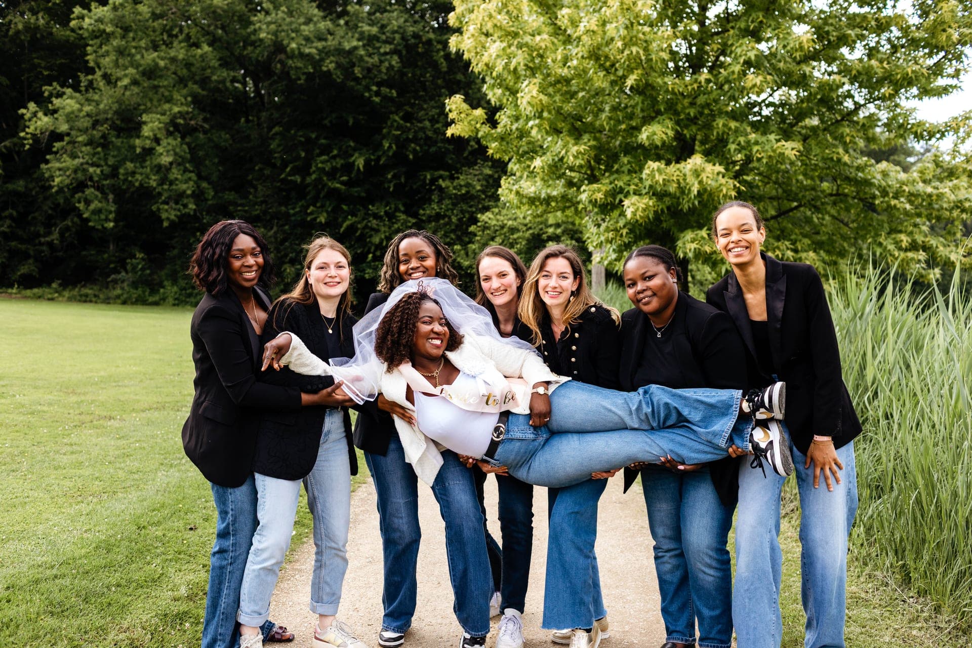 Les amies portent la future mariée voilée dans leurs bras éclat de rire sur un chemin de parc — reportage EVJF enterrement de vie de jeune fille Aurore Clercq Photographie Brabant Wallon