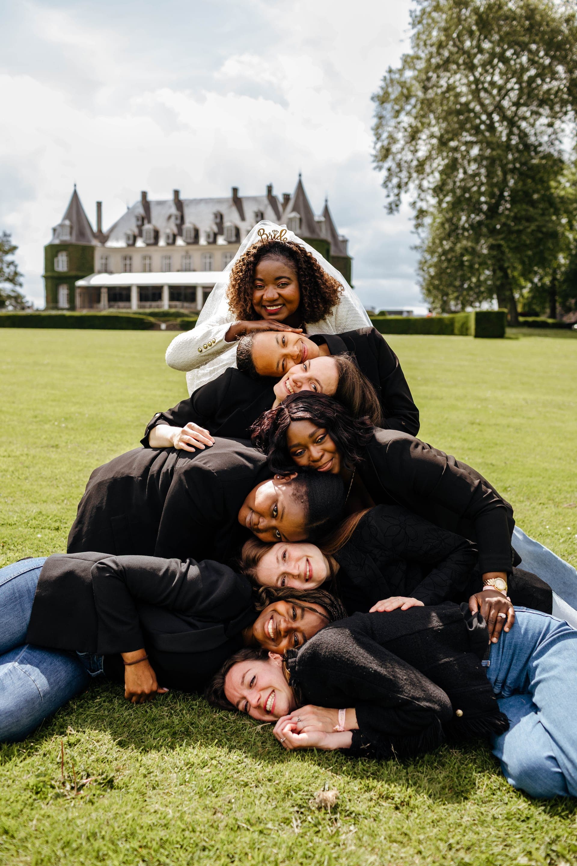 Amies allongées en pyramide sur la pelouse future mariée couronnée au sommet devant un château — reportage EVJF photo de groupe créative Aurore Clercq Photographie Belgique