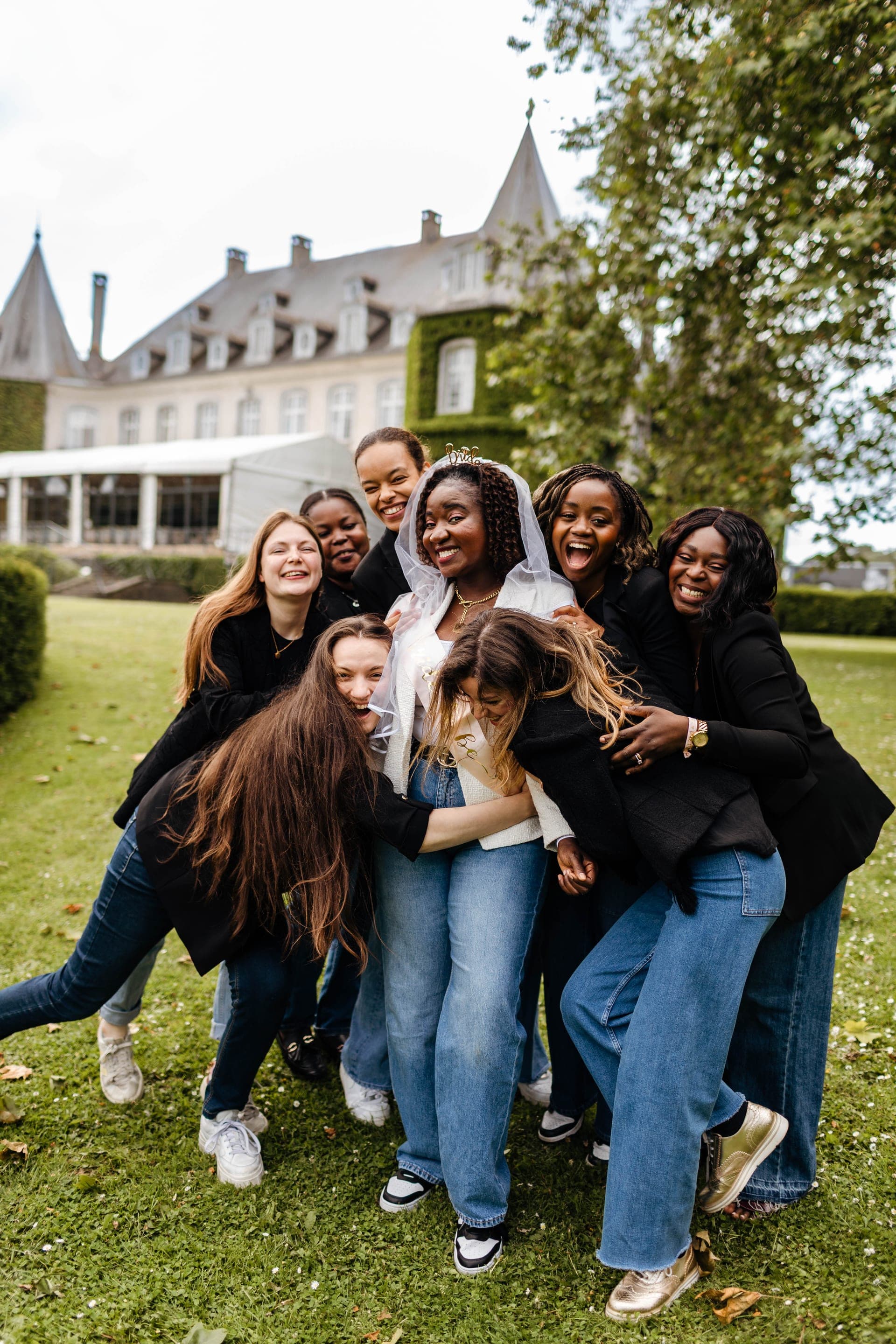 Groupe d'amies en câlin collectif fou rire autour de la future mariée voilée devant un château — reportage EVJF ambiance joyeuse Aurore Clercq Photographie Belgique