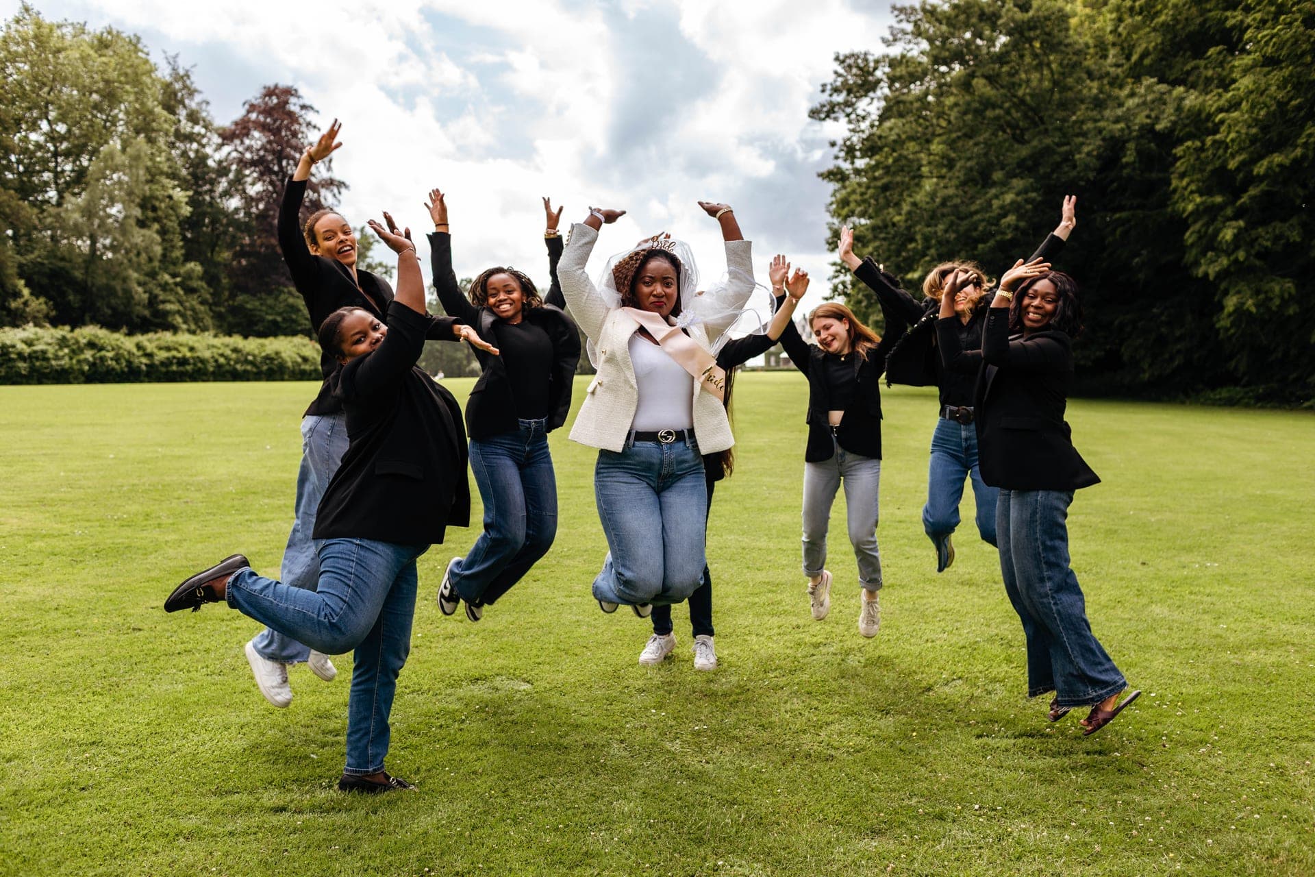 Groupe d'amies saute en l'air bras levés sur la pelouse autour de la future mariée énergie et joie — reportage EVJF ambiance Aurore Clercq Photographie Namur