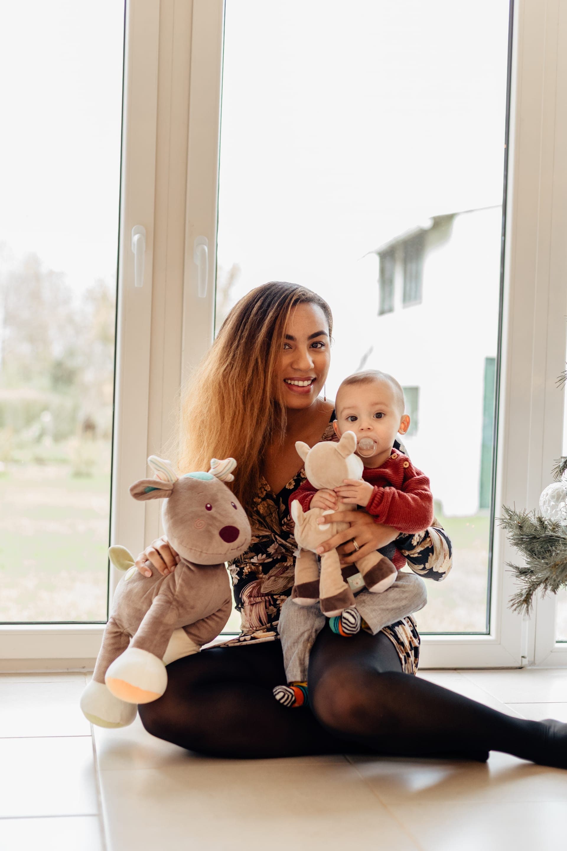 Maman et bébé avec peluches rennes devant la fenêtre — séance famille lifestyle Noël