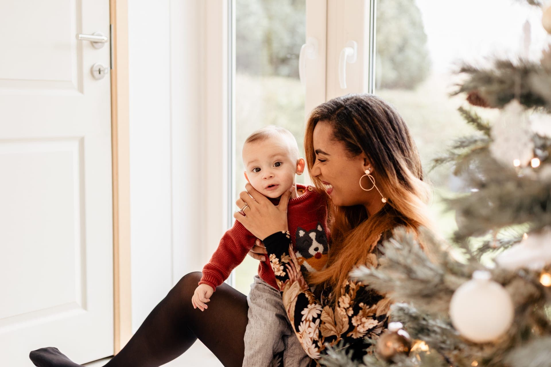 Maman et bébé près du sapin de Noël à la fenêtre — séance famille lumière douce