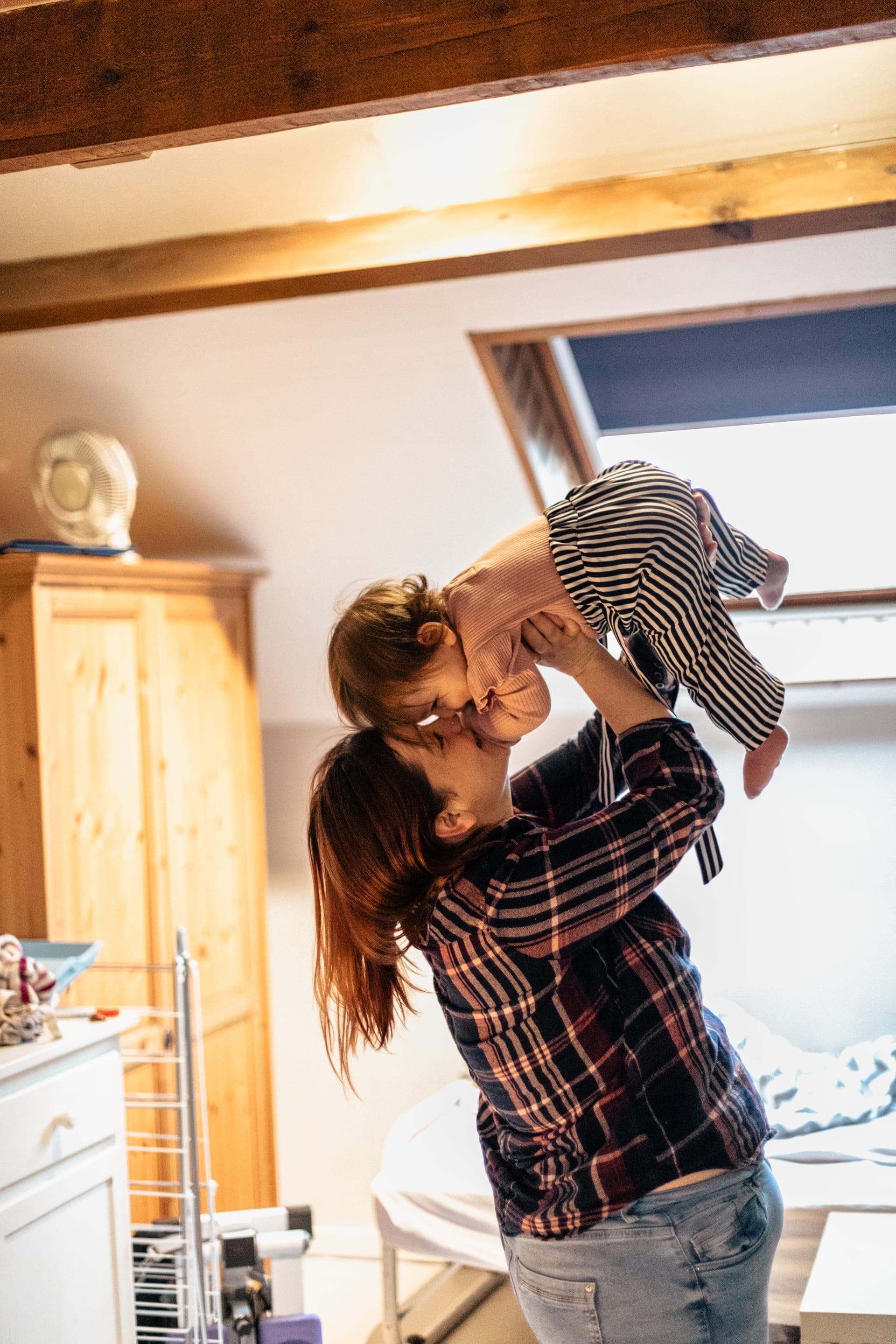 Maman soulève son bébé pour un bisou dans un grenier lumineux — séance famille tendresse