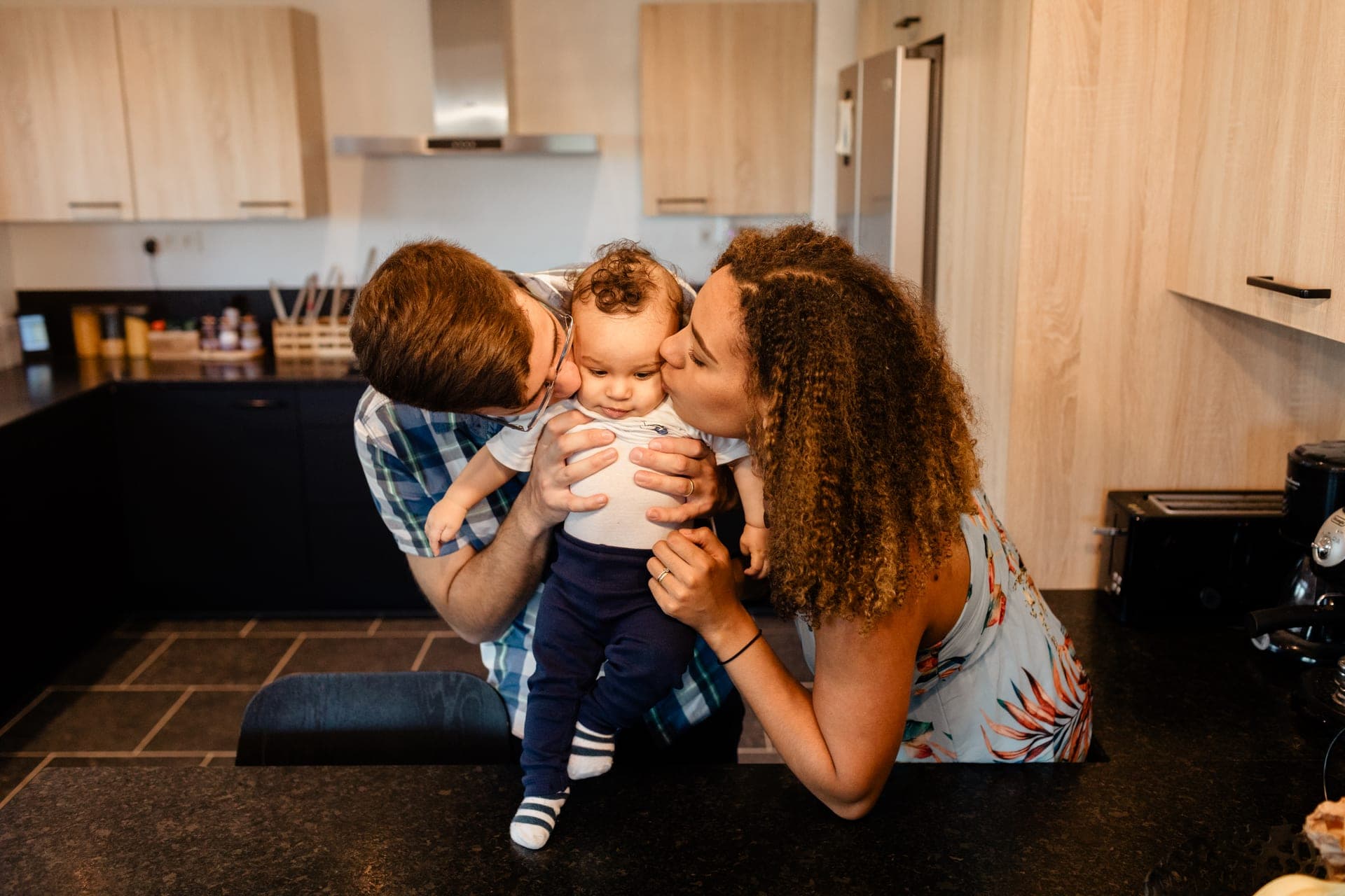 Parents embrassent leur bébé dans la cuisine — séance famille lifestyle Aurore Clercq Photographie Belgique
