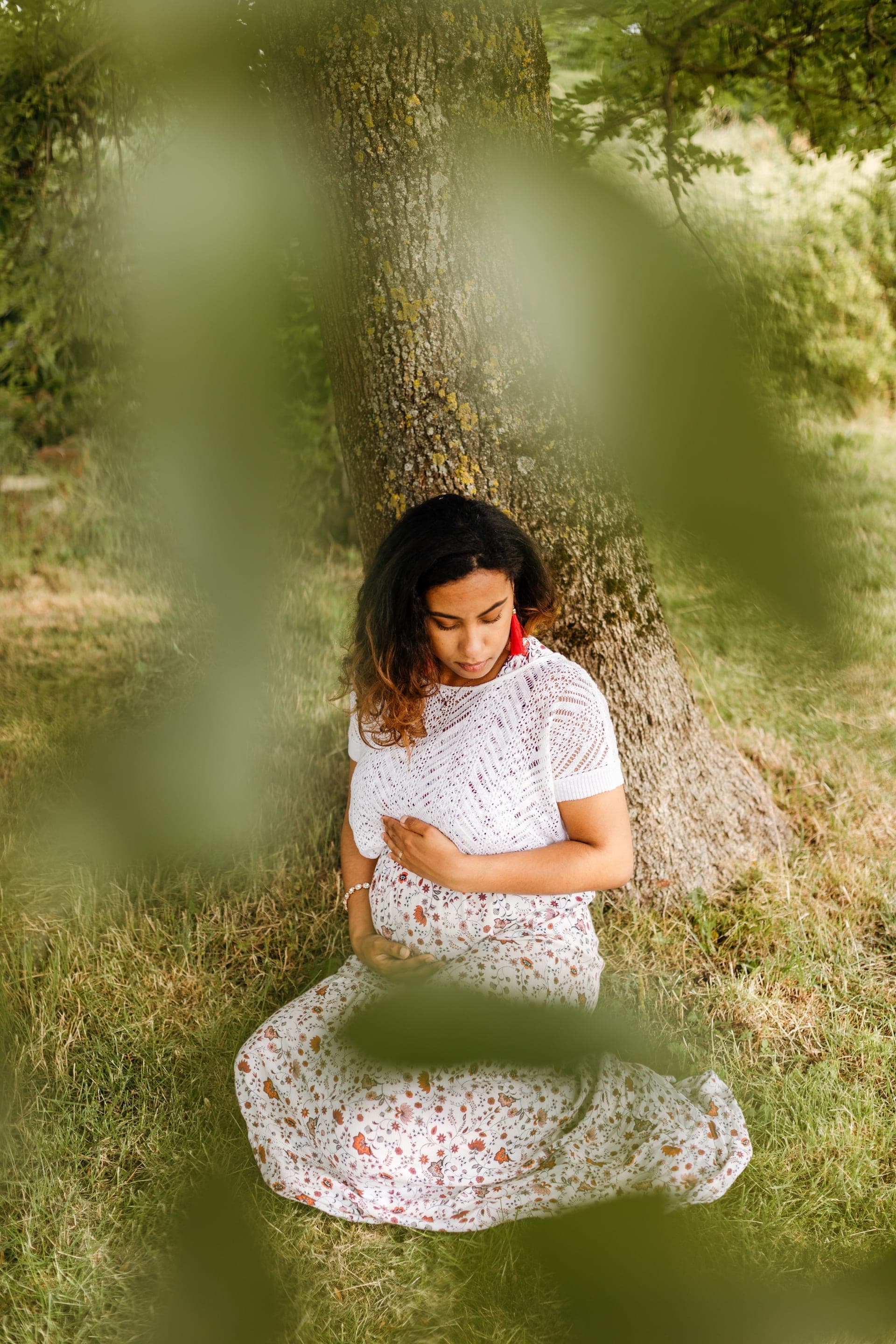 Future maman assise sous un arbre en robe blanche fleurie — séance grossesse nature