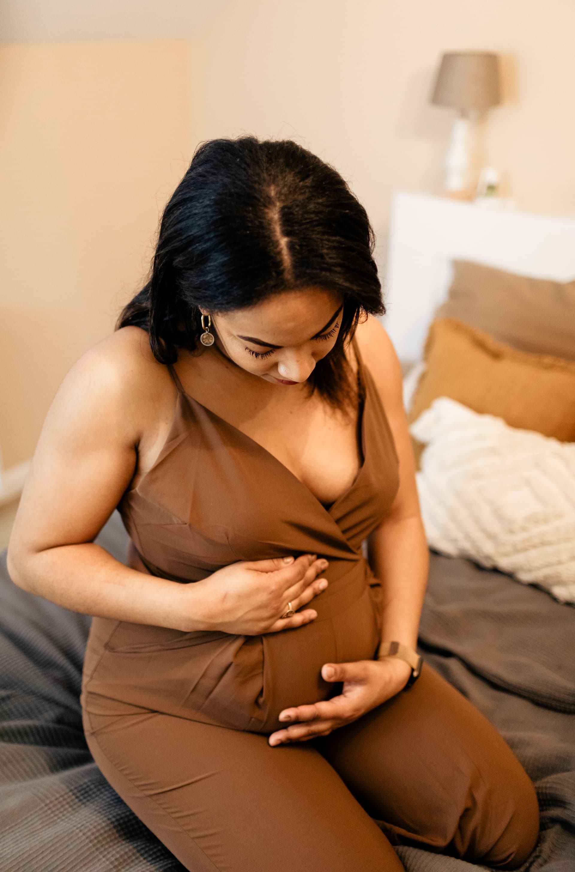 Future maman assise sur le lit robe café regard vers le ventre — séance grossesse douceur intimiste