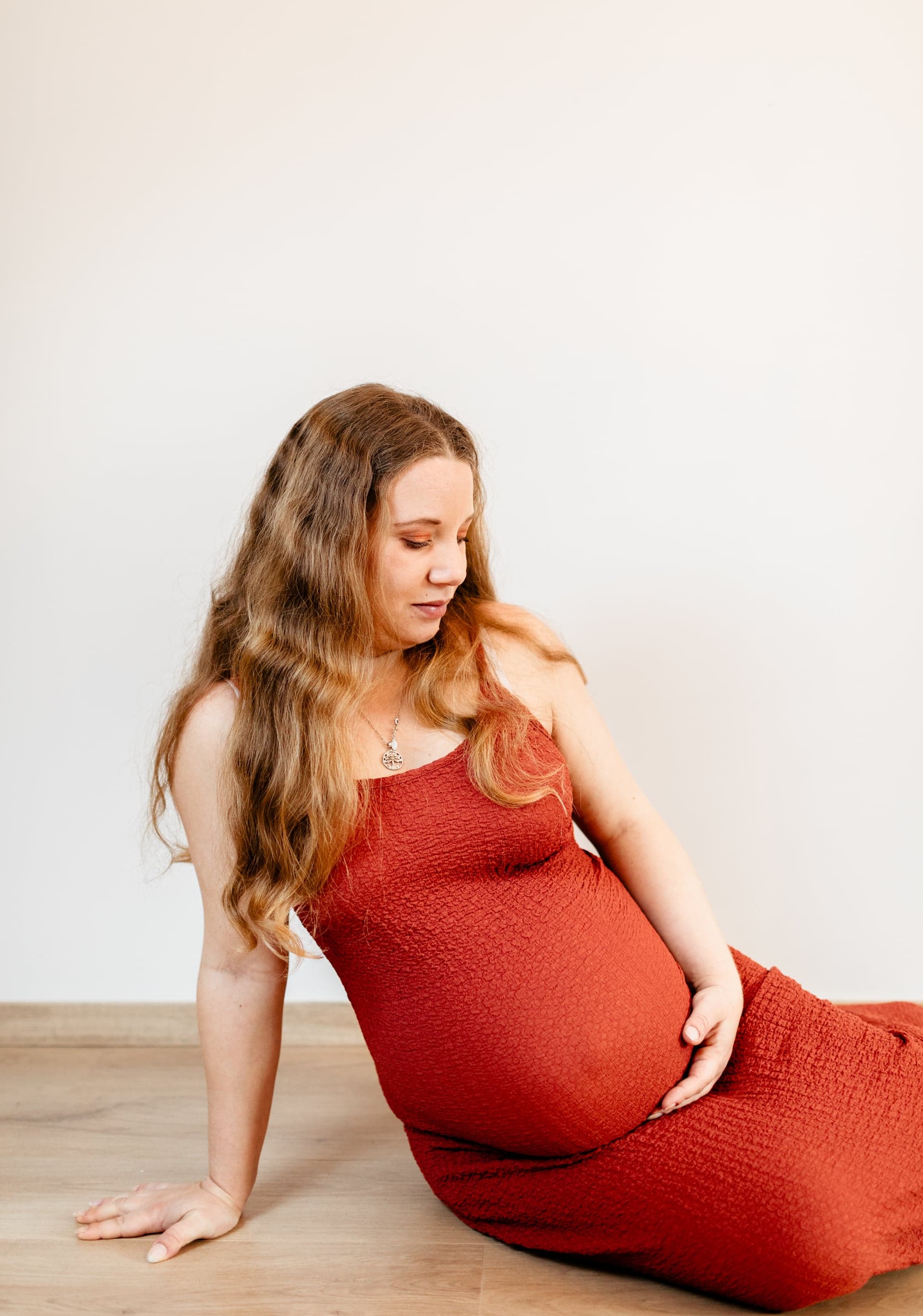 Future maman assise au sol en robe terracotta studio regard sur le ventre — douceur grossesse