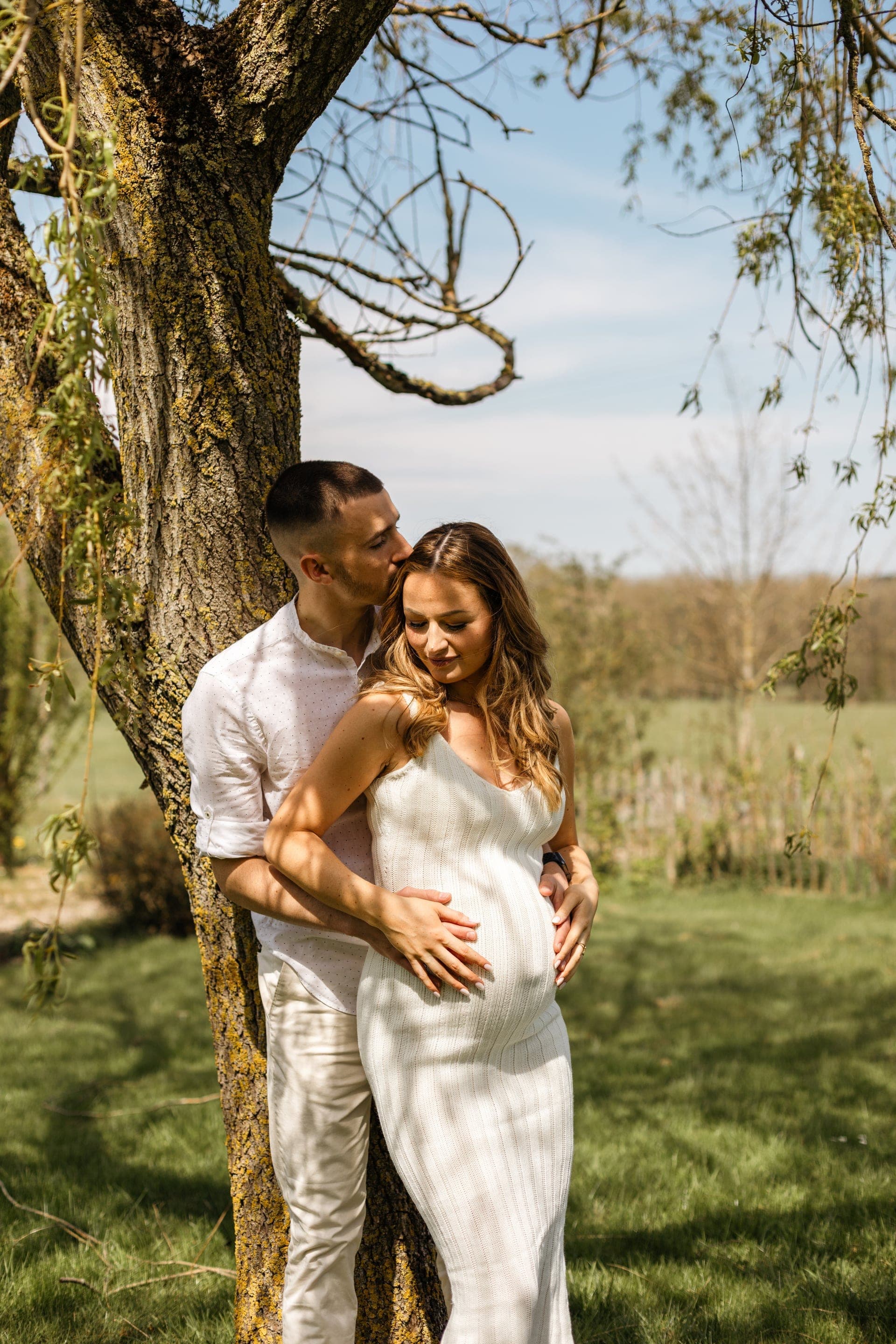 Couple enceinte enlacé contre un arbre en robe blanche baiser tendre — séance grossesse nature
