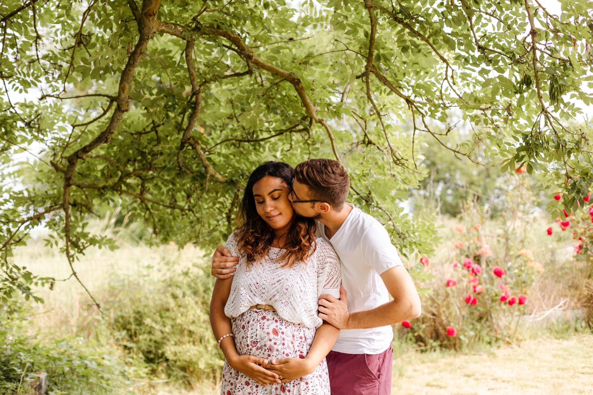 Couple enceinte baiser tendre sous arbre avec pivoines roses — grossesse poétique