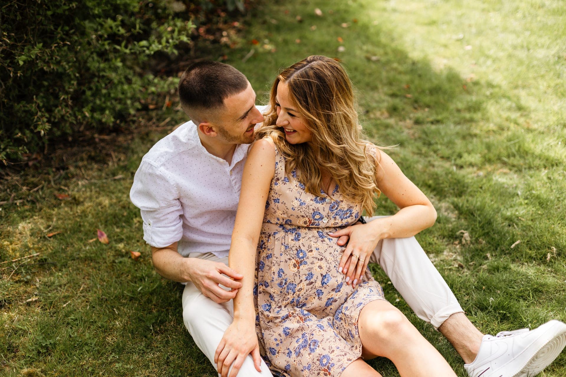 Couple enceinte assis dans l'herbe fou rire complicité — grossesse nature