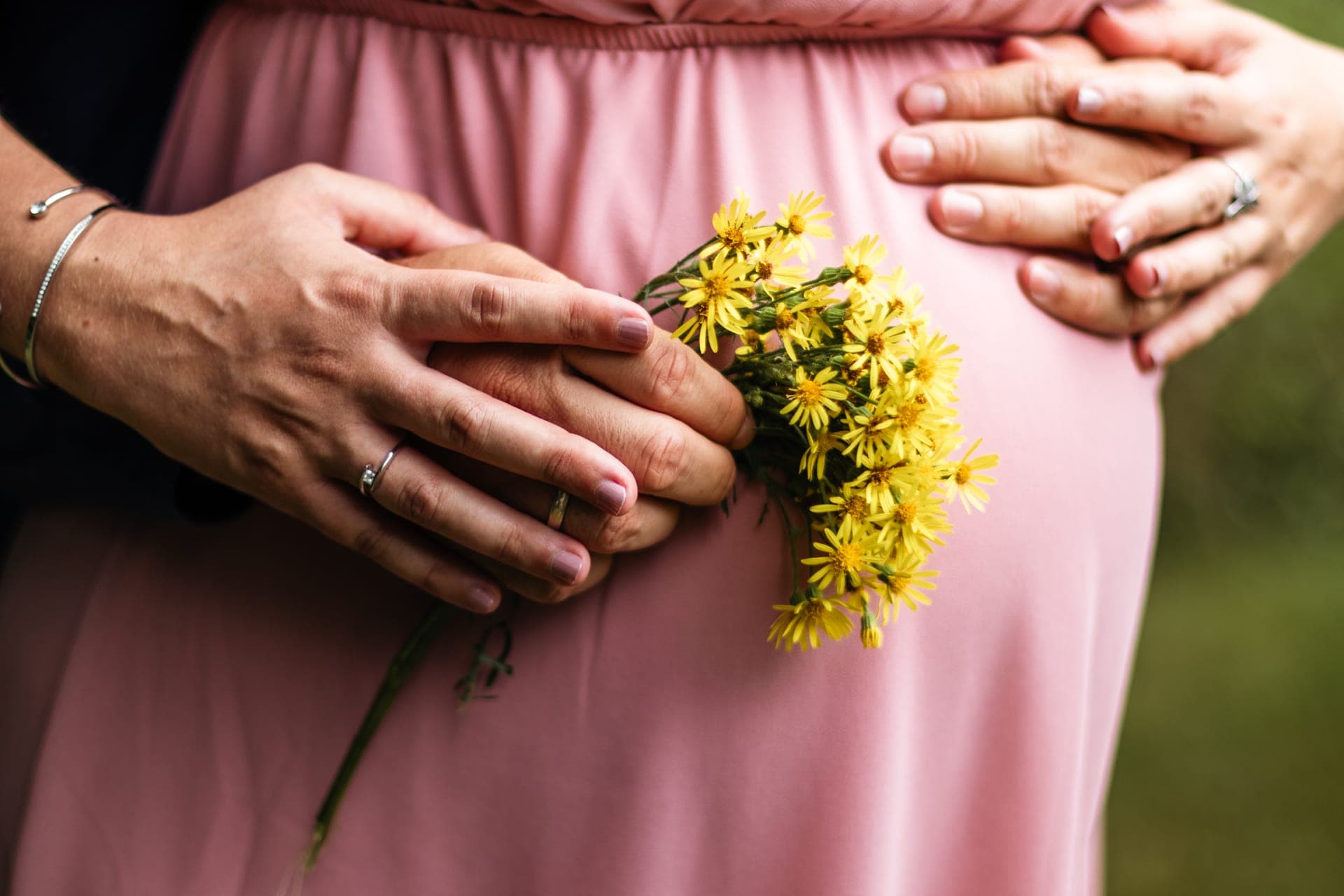 Mains du couple et bouquet de fleurs jaunes sur robe rose — détail grossesse