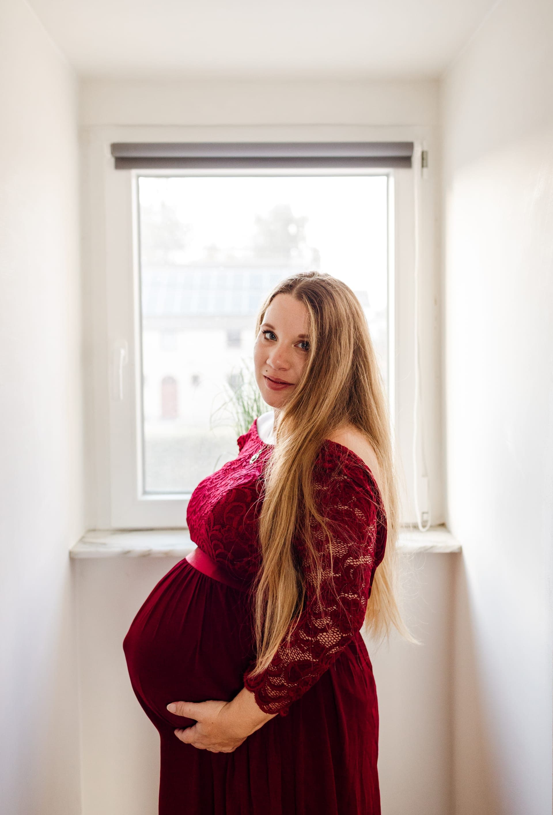 Future maman robe bordeaux dentelle près d'une fenêtre — lumière douce grossesse