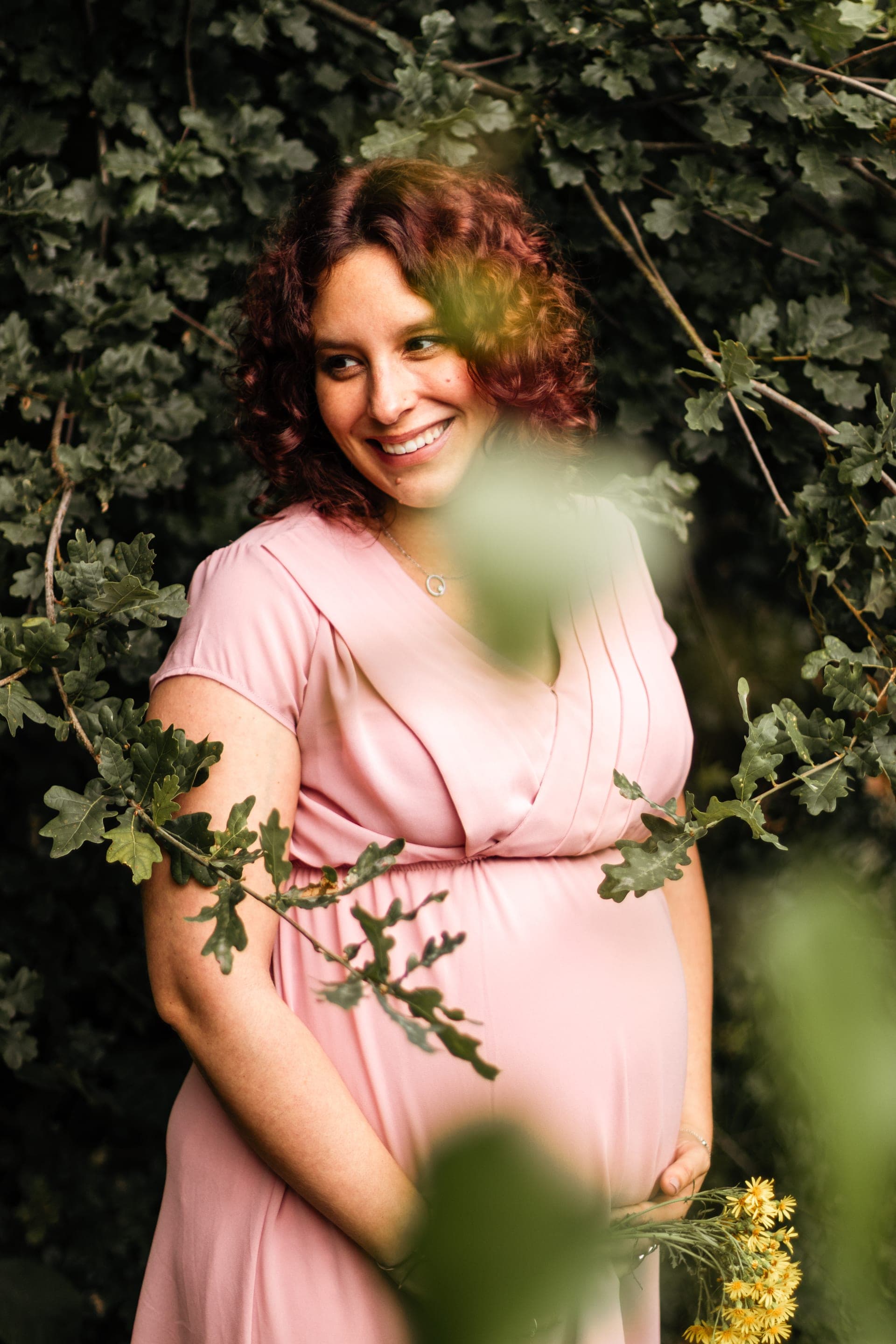 Future maman robe rose sourire radieux parmi les feuillages de chêne — séance grossesse nature
