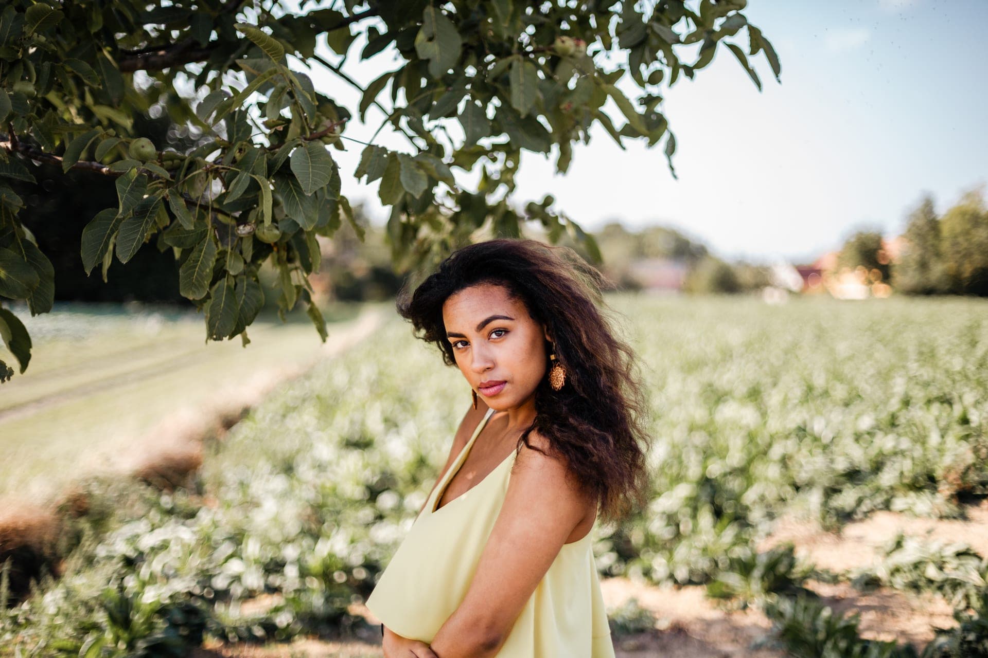 Portrait femme brune sous un arbre devant un champ vert top jaune regard intense — séance portrait été campagne