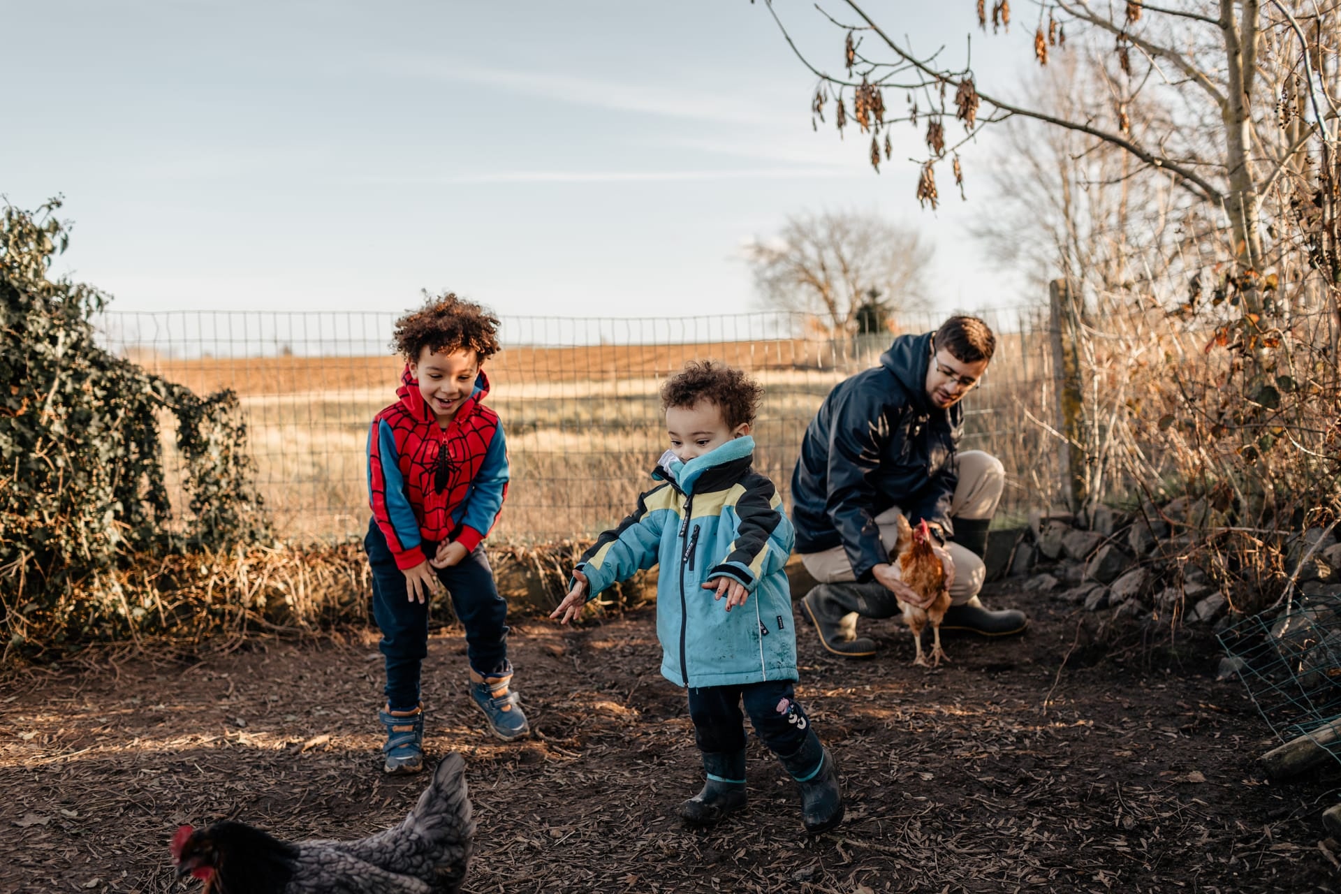 Papa et ses enfants observent les poules dans la cour d'une ferme à la campagne — séance photo famille en Brabant Wallon