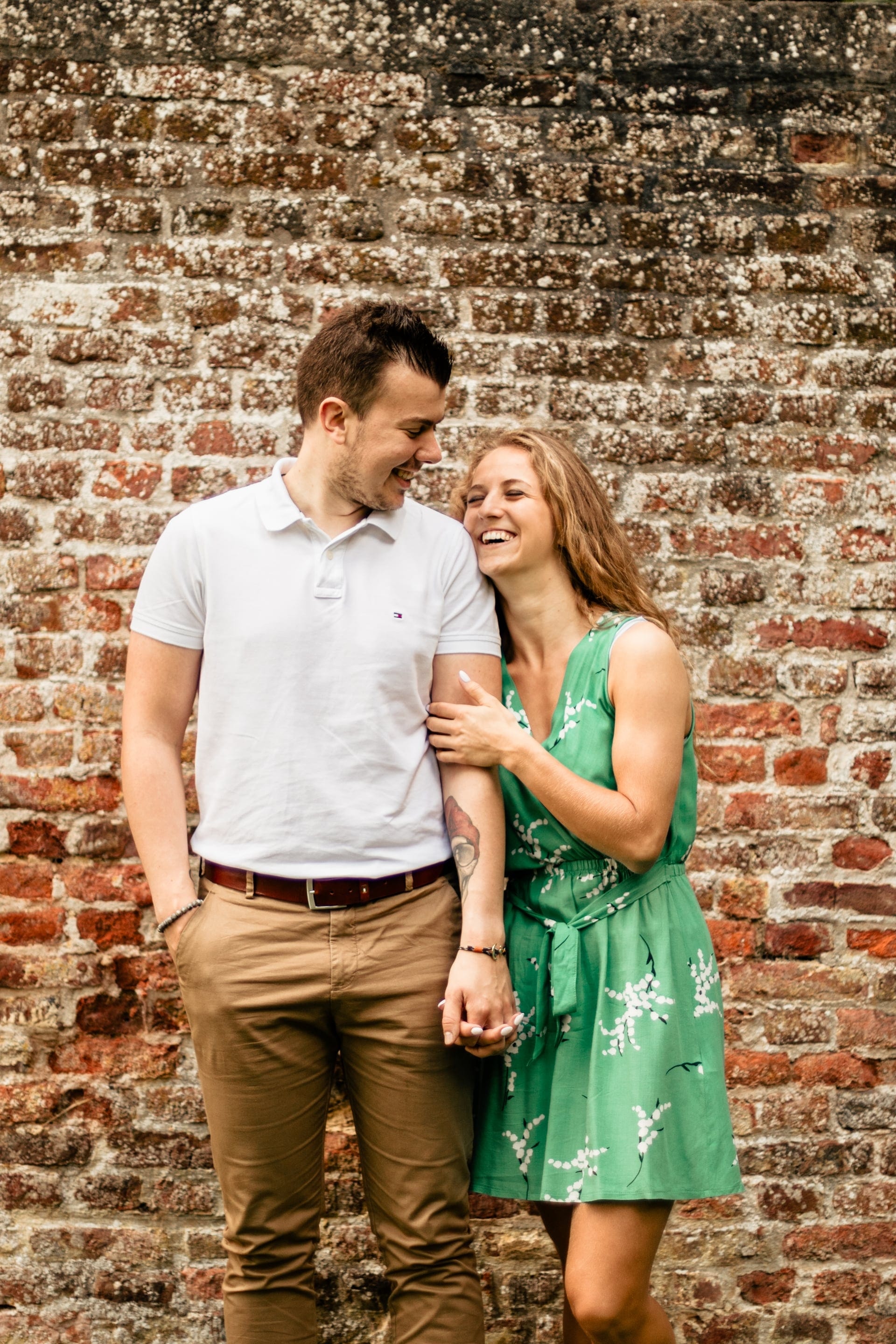 Couple en tenues harmonisées robe verte fleurie et polo blanc fou rire complice devant un mur de briques — exemple de tenues pour séance photo en Brabant Wallon