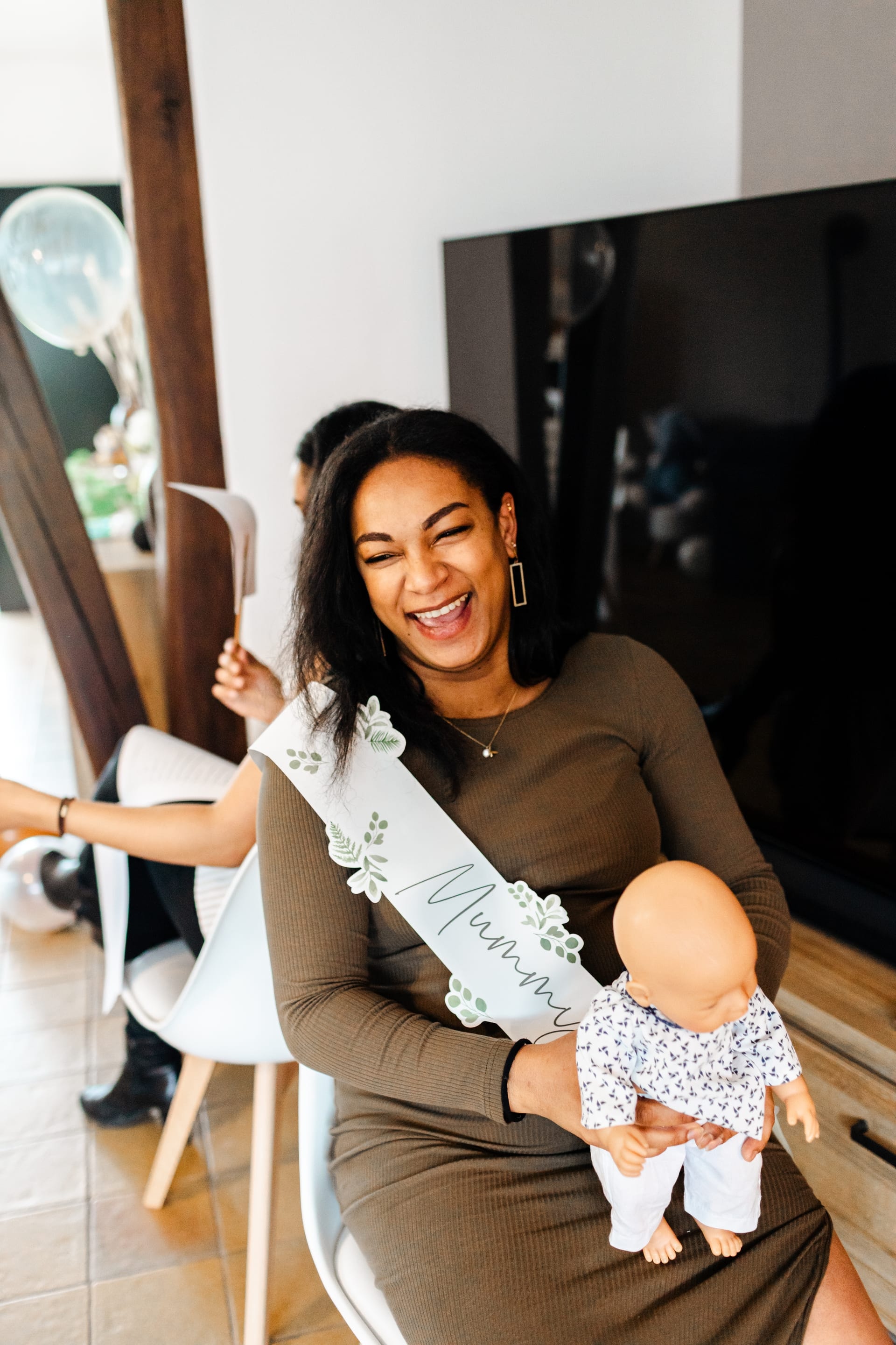 Future maman avec écharpe « Mummy to Be » et poupon, éclat de rire complice lors de sa baby shower — formule Reportage Lifestyle Aurore Clercq Photographie Brabant Wallon et Namur